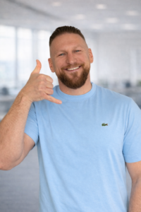 Mann in hellblauem T‑Shirt lächelt und macht eine Telefonhandgeste