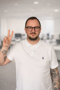 Mann mit Brille im weißen Poloshirt zeigt ein V-Zeichen; im Hintergrund ein unscharfes Büro